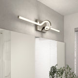 A modern wall-mounted LED light fixture with a looping design is installed above a large mirror. Light gray walls, a window with blinds, and a green plant are visible in a contemporary interior space.