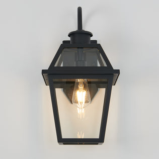 The Artika Colonial outdoor lantern wall light features a black metal frame with clear glass panels and an exposed incandescent bulb, mounted on a light-colored wall.