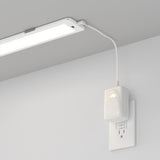 A rectangular LED light fixture is mounted under a shelf or cabinet, plugged into a white electrical outlet on the wall using a visible white power adapter and cord.