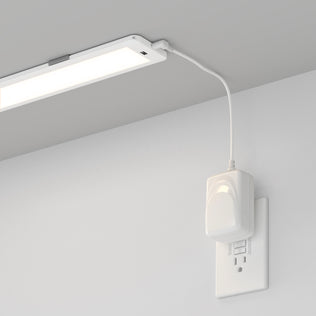 The Artika Maestro under cabinet LED light 3CCT is mounted beneath a gray shelf, with a white cord connecting it to a power adapter plugged into the wall outlet below.