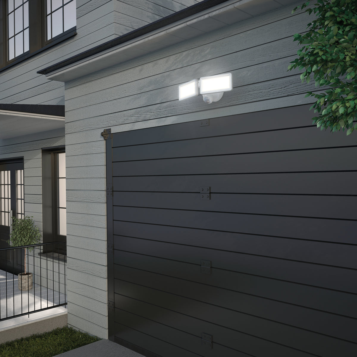A double floodlight is mounted above a modern dark gray garage door on a light-colored house exterior, illuminating the area at dusk near some foliage.