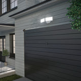 A double floodlight is mounted above a modern dark gray garage door on a light-colored house exterior, illuminating the area at dusk near some foliage.
