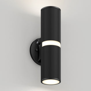 The Artika Groove LED indoor-outdoor wall light features a modern black cylindrical design with vertical ridges, emitting light from the top, bottom, and center band, and mounts on a round black base against a light wall.