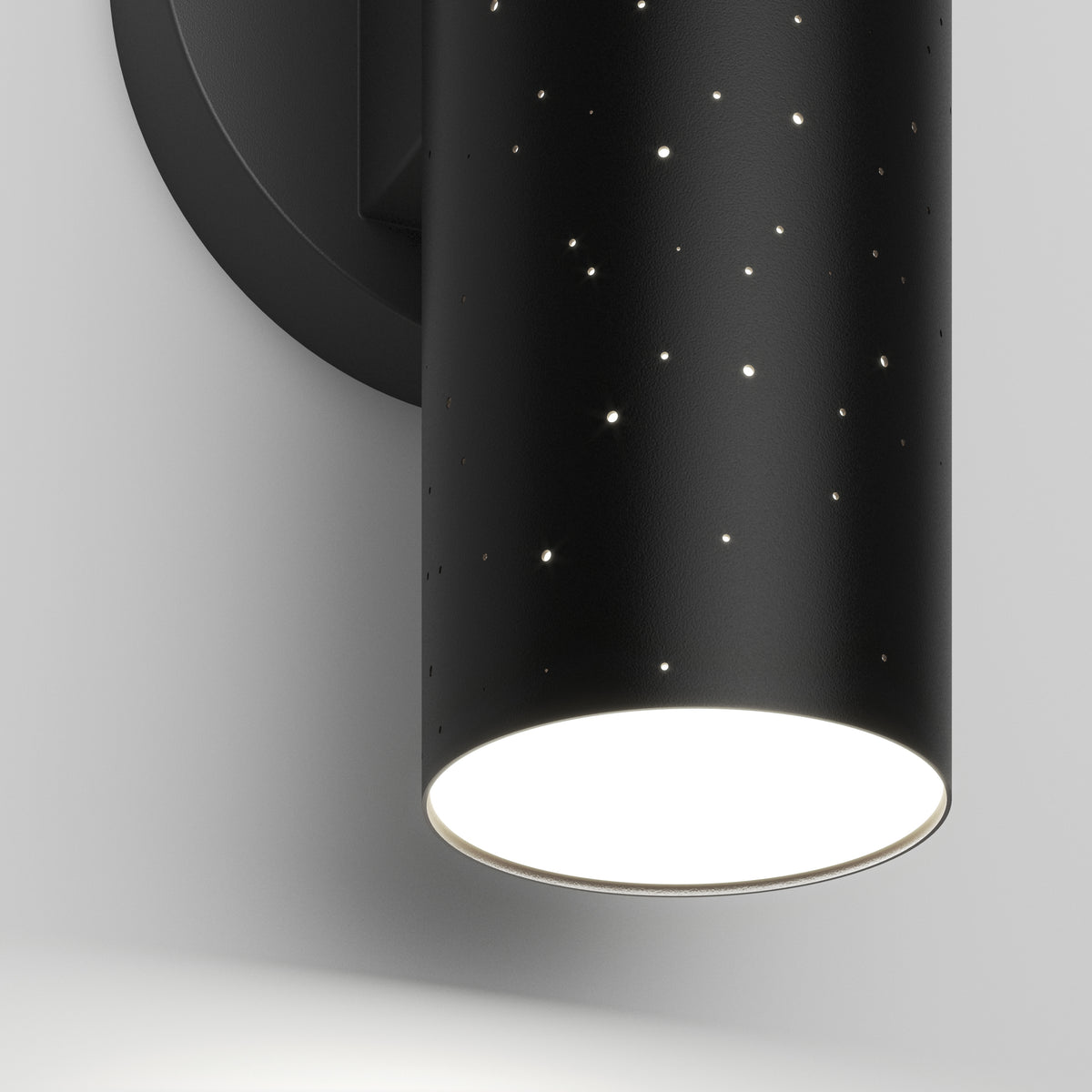 A close-up of a black cylindrical wall sconce with small star-like holes, emitting bright white light from the bottom. The fixture is mounted on a round black base against a white wall.