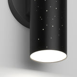 A close-up of a black cylindrical wall sconce with small star-like holes, emitting bright white light from the bottom. The fixture is mounted on a round black base against a white wall.