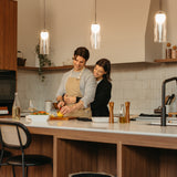 A couple stands in a modern kitchen, smiling as they prepare food together. The man, wearing an apron, slices a lemon while the woman hugs him from behind. Warm lighting and contemporary decor create a cozy atmosphere.