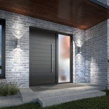 A modern house entrance with a dark horizontal-panel door, a tall frosted glass side panel, gray brick walls, two wall-mounted lights, and concrete steps with grass to the left.