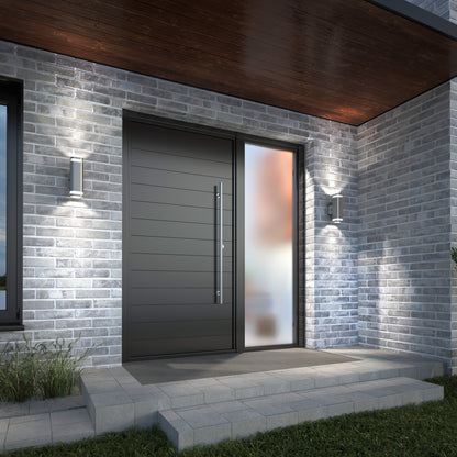 A modern front entrance featuring a dark sleek door, tall frosted glass side panel, brick walls, two Artika Mettle LED indoor-outdoor wall lights, and a small porch with concrete steps and grass on the side.