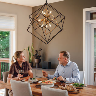 Two people dine and chat at a wooden table, illuminated by an Artika Imperium 9-light chandelier overhead.