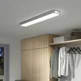 A modern rectangular ceiling light illuminates a closet area with wooden cabinets, a window, hanging clothes, and a shelf with storage boxes.