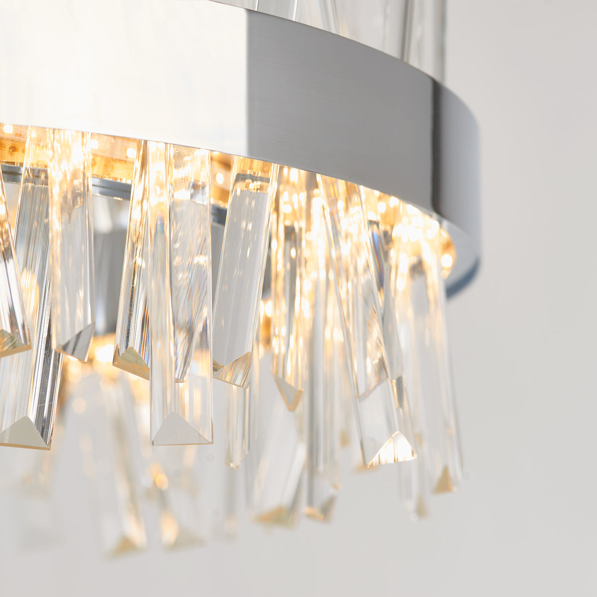 Close-up of a modern chandelier with clear, angular crystal prisms hanging from a circular metallic frame, illuminated with warm light against a neutral background.