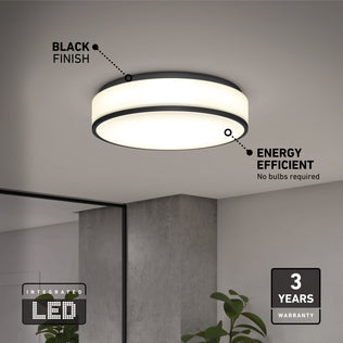 The Artika Giada 15 in. LED modern flush mount light (5 CCT), shown with a black finish in a minimal room, features integrated energy-efficient LEDs and a 3-year warranty. A plant and mirror are partially visible below the fixture.