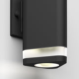 A close-up of a modern black wall-mounted outdoor light fixture with a cylindrical design and a frosted section near the top, emitting a soft white light downward.