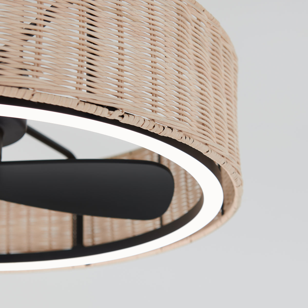 Close-up of a ceiling fan with black blades and a circular, wicker-patterned light fixture. The light glows around the edge of the woven shade, highlighting its texture against a light background.