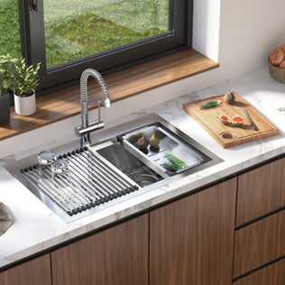 The Artika Newport double bowl stainless steel sink features a spring faucet, roll-up drying rack, and cutting board, set on a marble countertop with fresh produce, utensils, wooden cabinets, and a window overlooking greenery.