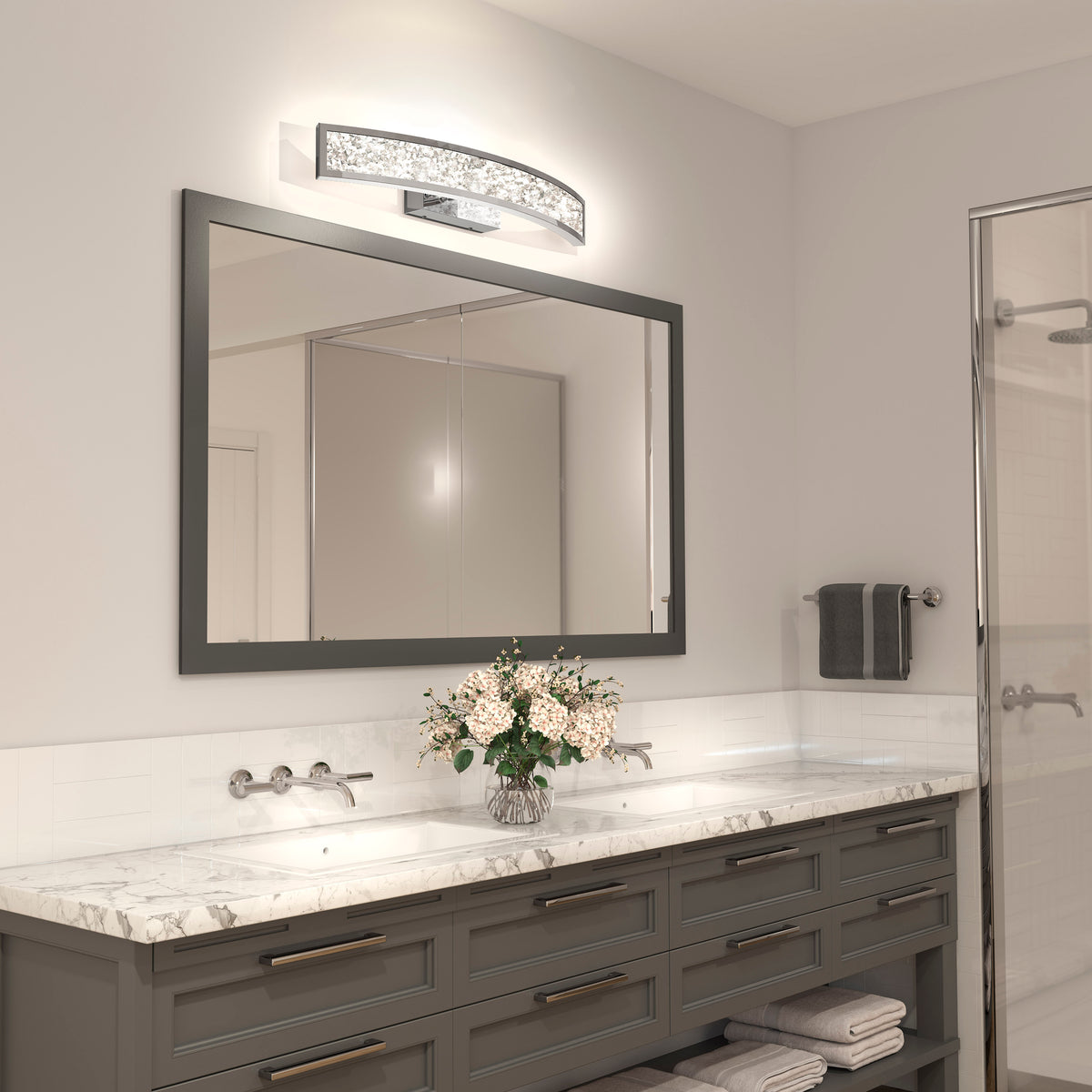Modern bathroom with a large mirror, marble countertop, double sink with silver fixtures, gray cabinets, flowers, towels, and the Artika Glitter 27-in Integrated LED Vanity Light Bar. Glass shower is visible on the right.