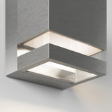 A close-up view of a modern, square brushed metal wall light fixture with a frosted glass bottom, emitting soft white light onto a light-colored wall.