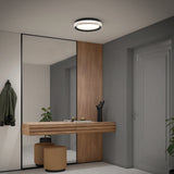 A modern hallway with a large mirror, wooden floating console, two round tan stools, green coat on a hook, potted plant, decorative items, gray door, and a circular ceiling light.