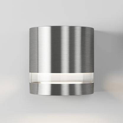 The Artika Valor Round LED indoor-outdoor wall light features a modern cylindrical design in brushed metal with a horizontal frosted section that emits a soft glow, shown mounted on a plain white wall.