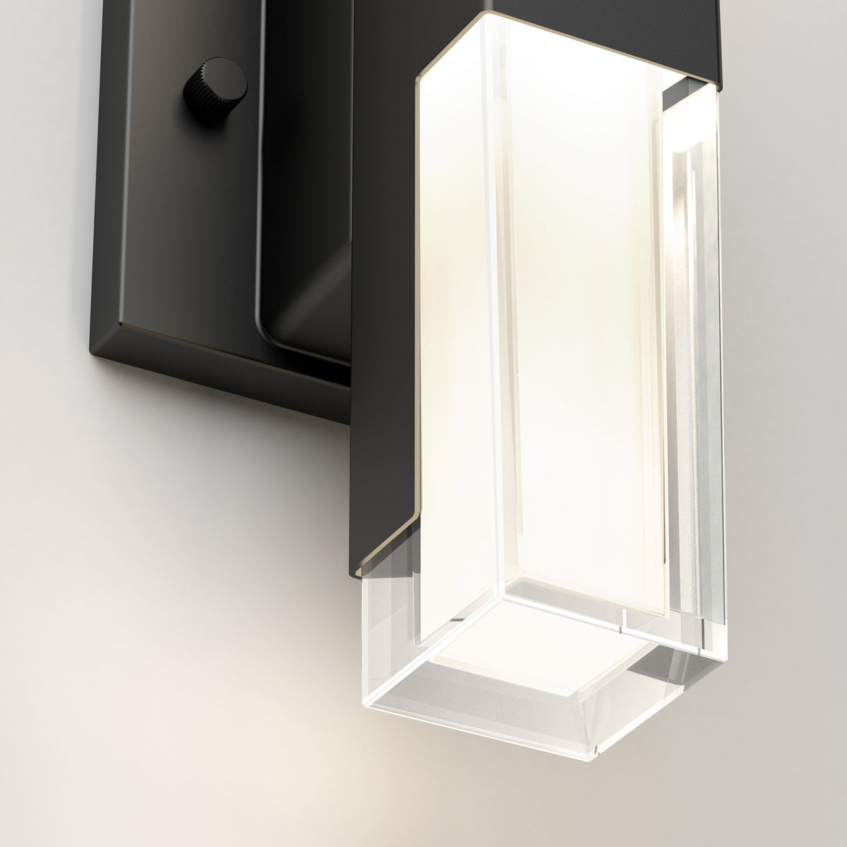 A close-up of a modern wall-mounted light fixture with a rectangular black base and a clear glass shade, illuminating a soft white light against a neutral wall.