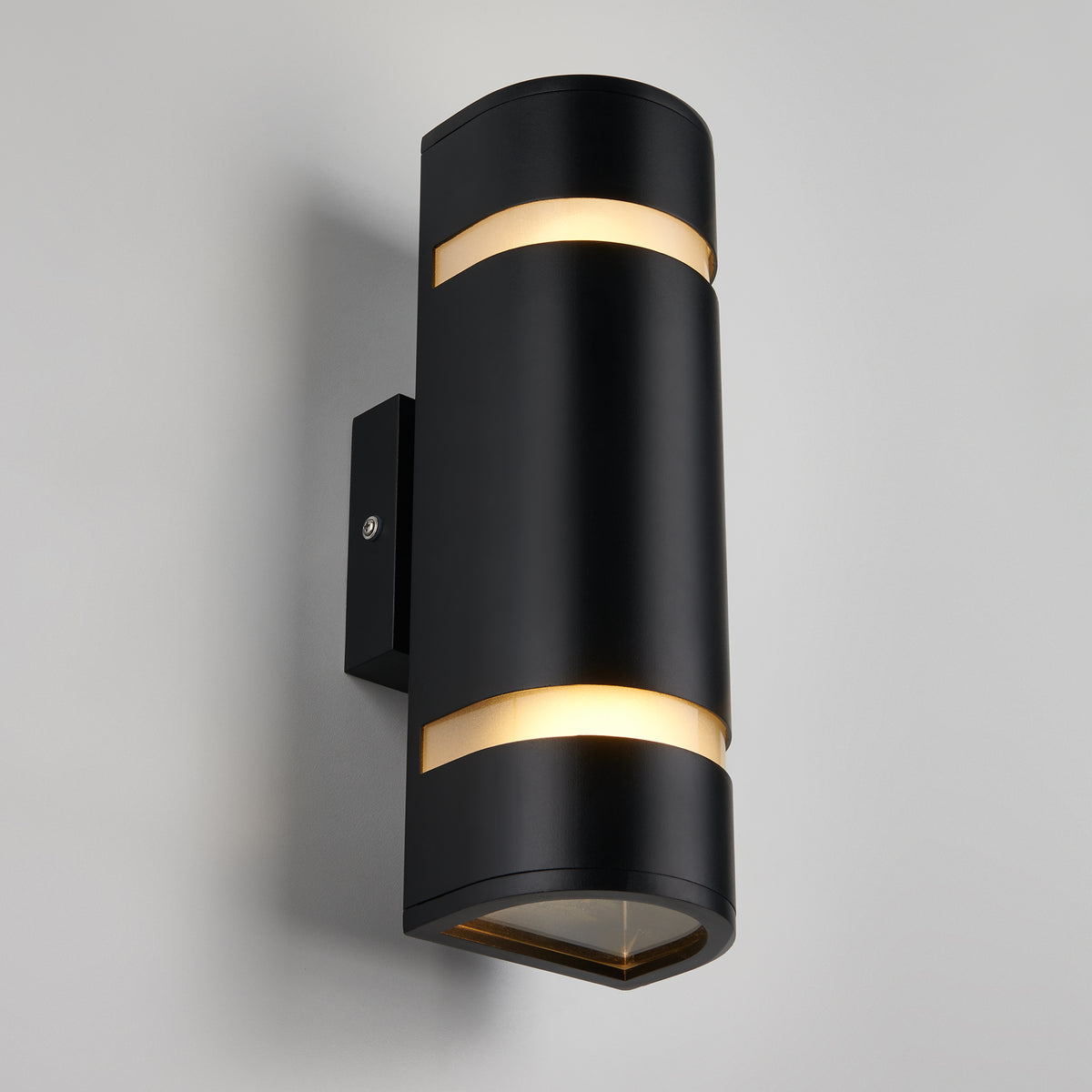 A modern black wall sconce with a cylindrical shape, featuring two horizontal slits near the top and bottom that emit soft, warm white light, mounted on a light gray wall.
