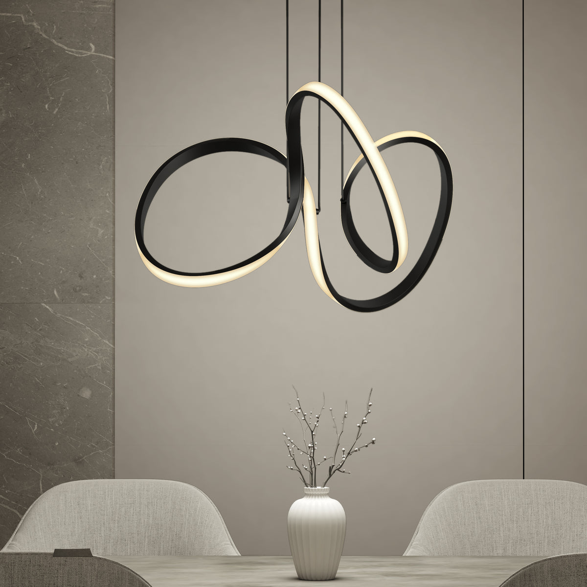 A modern dining area with a sculptural, loop-shaped black and gold pendant light above a table. On the table is a white vase containing minimalist branches. Two light-colored chairs are partially visible.