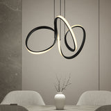 A modern dining area with a sculptural, loop-shaped black and gold pendant light above a table. On the table is a white vase containing minimalist branches. Two light-colored chairs are partially visible.
