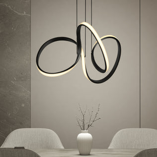 A modern dining room features the Swirl LED pendant light by Artika in black and gold above a table with a white vase and minimalist branches, flanked by two light chairs against neutral walls.