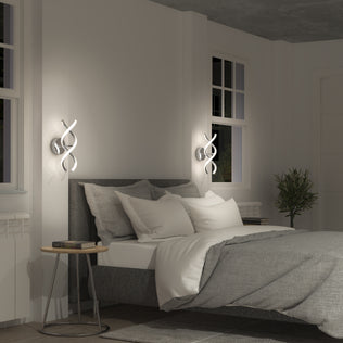 A modern bedroom with a gray upholstered bed, white and gray bedding, round wooden side table, Artika's DNA modern LED wall sconce 3 CCT, a window with white trim, and a potted plant in the corner.