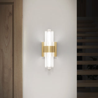 The Artika Dorado contemporary LED wall sconce 3 CCT features vertical glass rods and a gold metallic center, mounted on a white wall by a wooden door and stairs. This luxury fixture provides soft, diffused illumination.