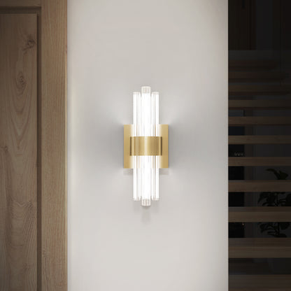 The Artika Dorado LED contemporary wall sconce 3 CCT features a gold metallic center and vertical glass rods, emitting soft light while mounted on a white wall beside a wooden door and open staircase.