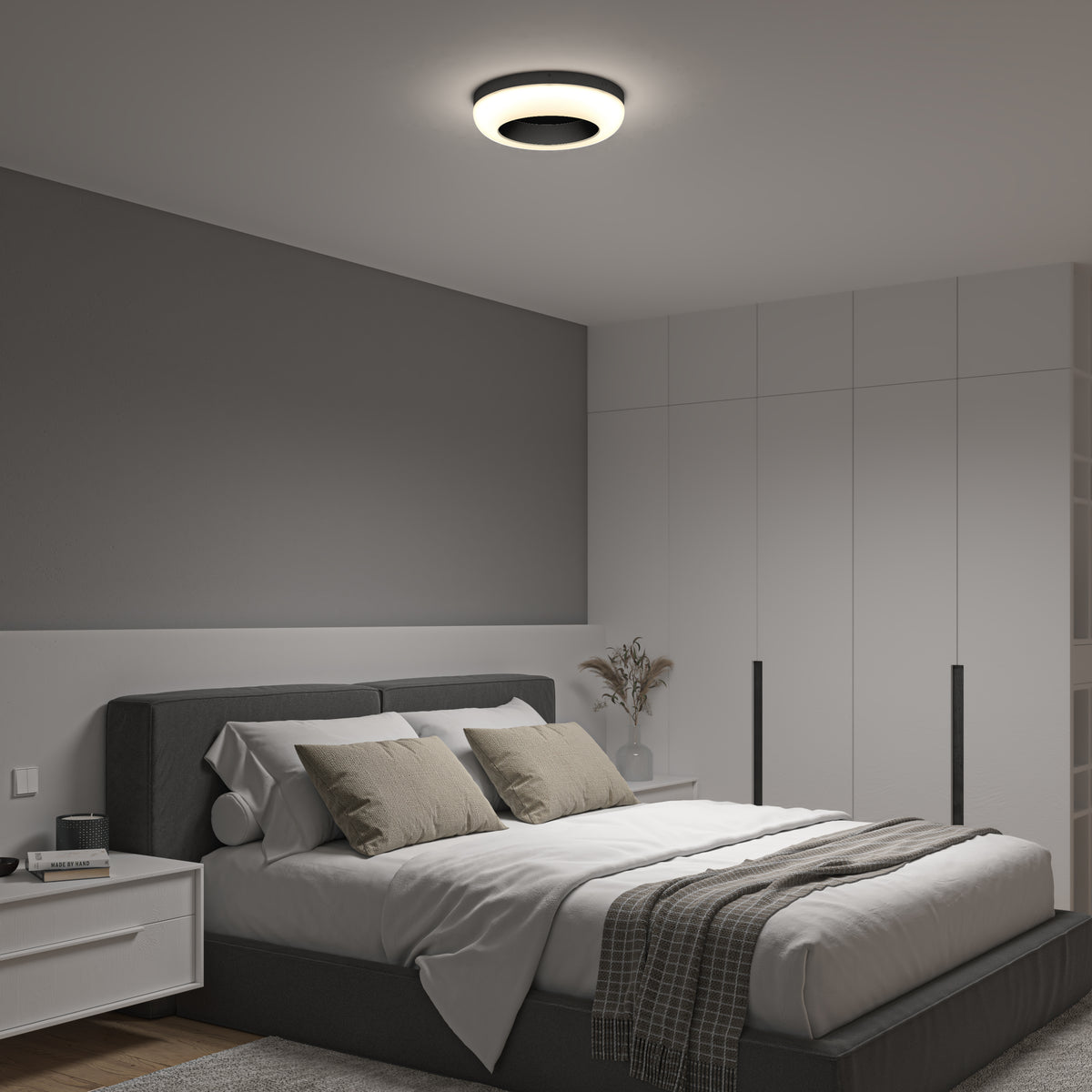 Modern bedroom with a gray accent wall, neatly made bed with neutral bedding and pillows, a round ceiling light, white built-in wardrobes, a bedside table, and a vase with dried flowers.