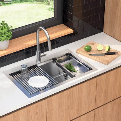 The Artika Newport single bowl stainless steel workstation sink features a drying rack holding a glass, plate, and herbs. On the counter is a cutting board with knife and limes. A potted plant sits on the windowsill overlooking greenery.