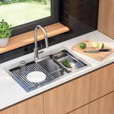 A modern kitchen sink with a faucet, dish rack, glass, white plate, and herbs in the sink. On the counter, there’s a wooden cutting board with a knife, a halved lime, and a whole lime. Greenery is outside the window.