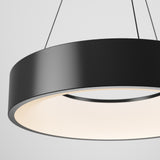 A close-up view of a modern, circular black pendant light fixture with a smooth finish, suspended by thin cables against a plain white background. The light glows softly from the inner edge.