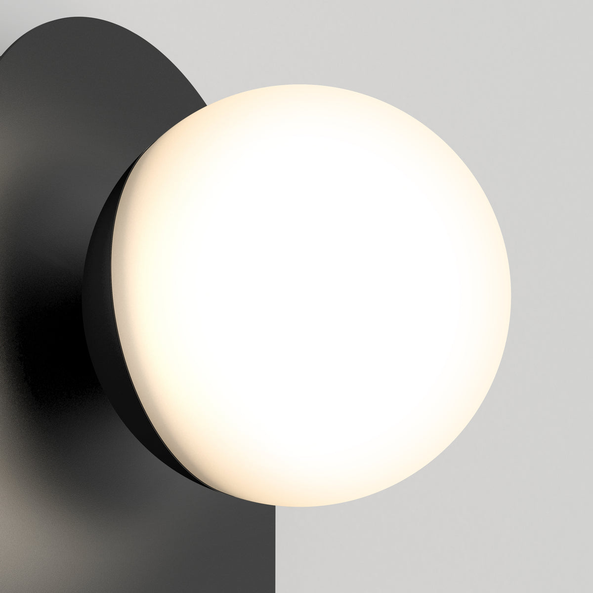A modern wall sconce with a round, frosted white glass shade and a black metal base, mounted on a light gray wall. The light emits a soft, warm glow.