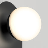 A modern wall sconce with a round, frosted white glass shade and a black metal base, mounted on a light gray wall. The light emits a soft, warm glow.