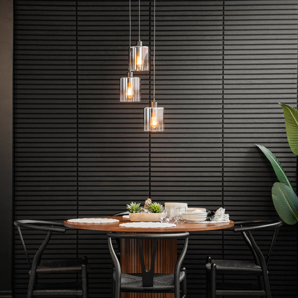 A modern dining area featuring a round wooden table for four, black chairs, a Haze modern 3-light glass pendant by Artika above, a textured black wall, and a large green plant on the right.
