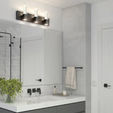 Modern bathroom with a large mirror, three-bulb vanity light, black fixtures, a white countertop, a potted plant, a towel hanging on a rack, and a glass shower enclosure reflected in the mirror.