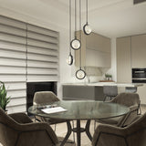 Modern kitchen and dining area with round glass table, four upholstered chairs, hanging pendant lights, neutral cabinetry, marble backsplash, a plant, and large window with striped roller blinds.