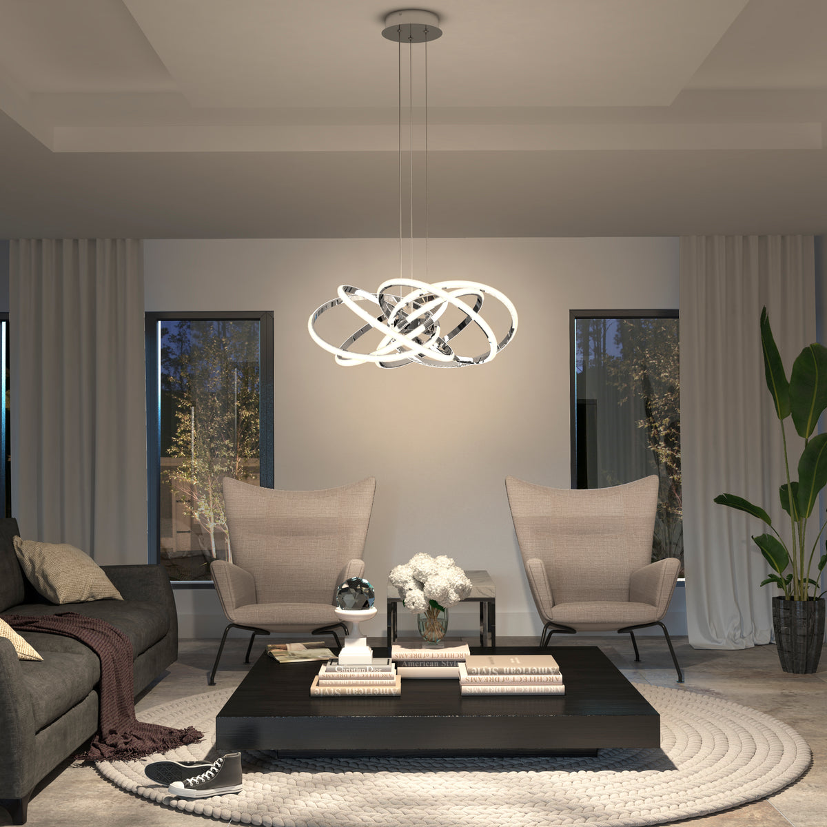 Modern living room featuring two beige armchairs, a dark sofa, black coffee table, books, flowers, large windows with white curtains, a tall plant in the corner, and the Artika Maelstrom modern LED pendant light 5 CCT.