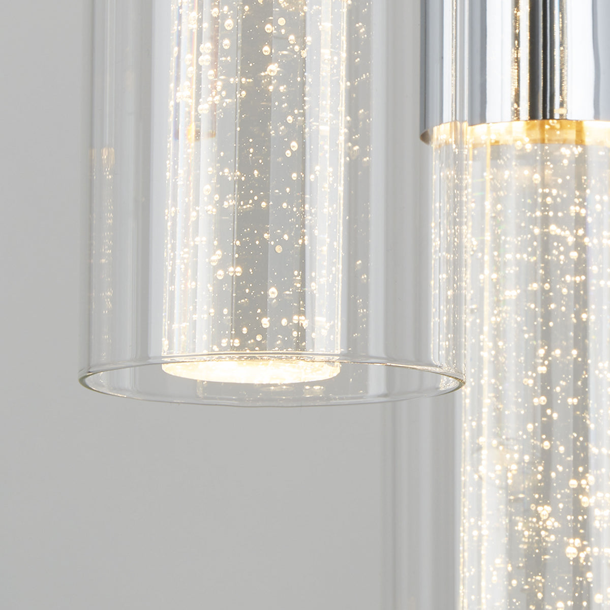 Close-up of a modern glass pendant light with bubbles inside the cylindrical glass shade, illuminated and hanging against a plain, light gray background.