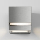 A modern, square-shaped wall light fixture made of brushed metal, featuring a minimalist design with a soft, diffused light glowing from the top and bottom edges against a plain white wall.