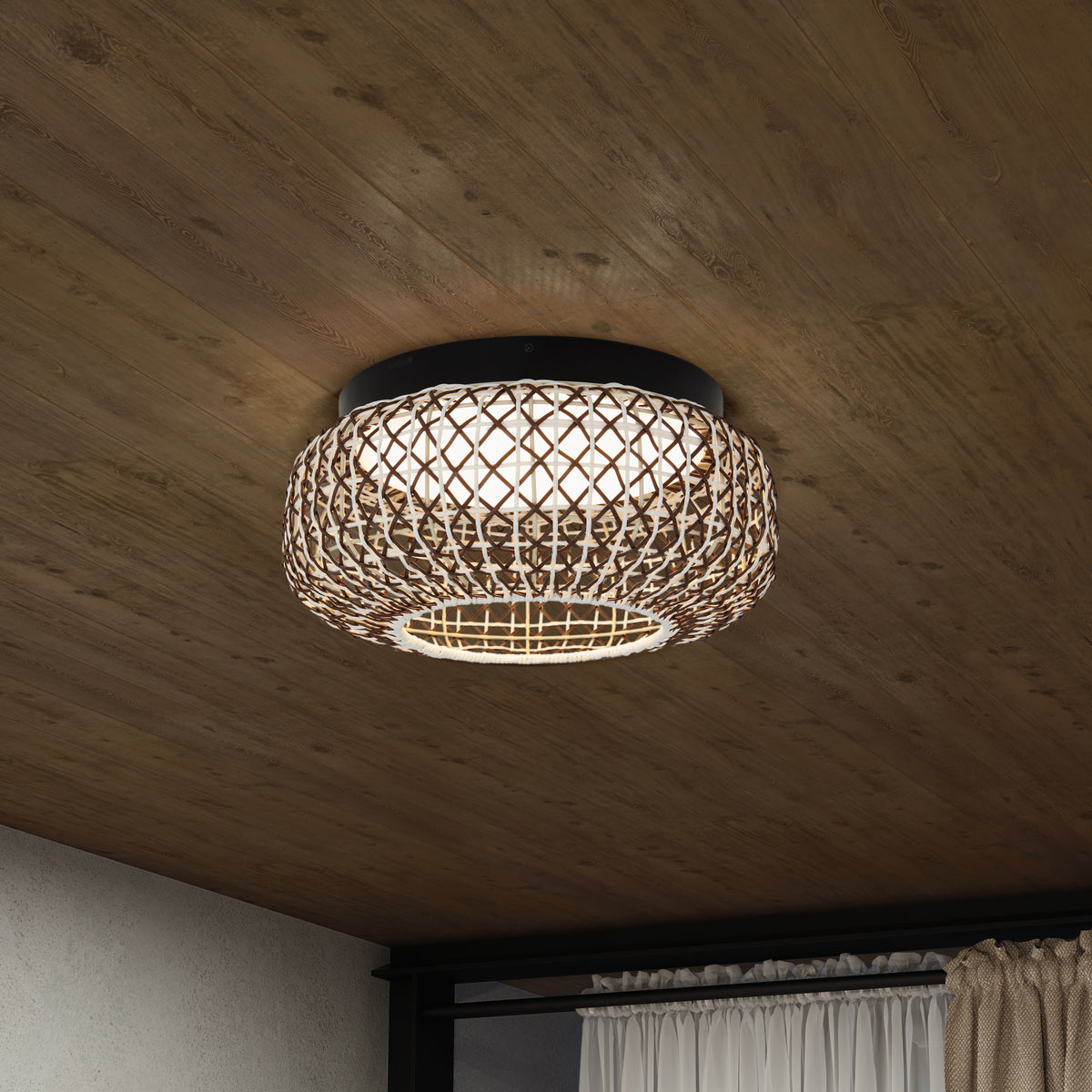 A woven rattan ceiling light fixture with a round shape is mounted on a wooden ceiling, illuminating a cozy interior space with curtains visible in the background.