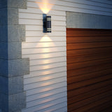 A modern outdoor wall light is mounted on beige siding next to a wooden garage door, illuminating the wall with beams of light both upward and downward.