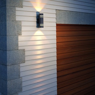 The Artika V3 indoor-outdoor wall light is mounted on white siding near a wooden garage door, casting warm up and down illumination on the wall.