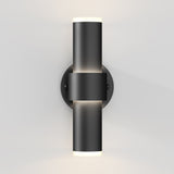 A modern black wall sconce light fixture with a cylindrical design, mounted on a round base, emitting light from both the top and bottom ends against a white wall.
