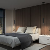 A modern bedroom with a bed featuring white and gray bedding, a dark wood accent wall, two pendant lights, white nightstands with small plants, and a city view through the window at night.