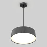 A modern pendant ceiling light with a round, ribbed, black lampshade hanging from a black rod, glowing brightly against a plain light gray background.