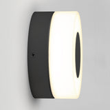 A modern, oval-shaped wall sconce with a black base and a glowing white frosted cover, mounted on a plain light gray wall.
